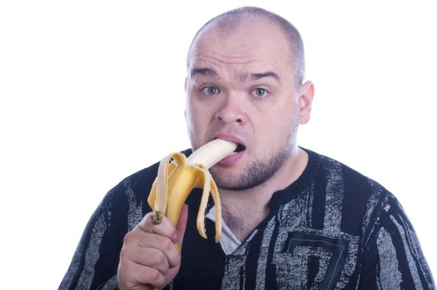 Young bald serious man eats yellow banana